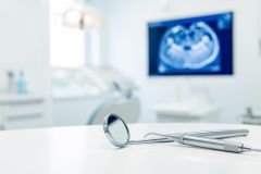 Professional dental instruments, including a silver mirror and handpiece, sit on a white table in a bright dental office. The blurred background features a medical monitor displaying a blue X-ray scan and a dental chair, highlighting a modern, sterile clinical setting for oral healthcare and dental examinations.
