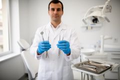 Dentist in a bright exam room wearing blue gloves holding stainless dental instruments toward the viewer.