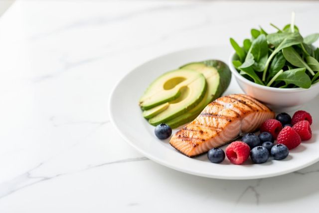 A nutritious meal featuring a grilled salmon fillet, creamy avocado slices, fresh blueberries, and red raspberries on a white plate. A side of green leafy salad sits in a small bowl. Set on a white marble tabletop, this colorful and healthy dish is ideal for keto or low-carb diets.