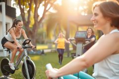 Woman cycling outdoors on a stationary bike. Two other women are exercising nearby. Sunny day, green grass, and a vibrant outdoor fitness environment. Active lifestyle, workout, and healthy living theme.
