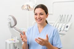 A smiling female dental professional wearing light blue scrubs in a bright, modern dental clinic. She is gesturing with her hands while talking in a clean, clinical environment. The background includes a dental chair, medical exam light, and professional dental instruments, creating a professional healthcare atmosphere.