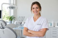 Smiling dental clinician standing in a bright clinic with chair and instruments visible.