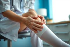 Doctor examining patient's leg, wearing a white coat and socks. Medical examination setting with blue background and equipment visible. Focus on leg and diagnostic assessment.