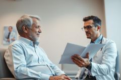 Doctor consults with a senior patient in a modern clinic. The image shows a healthcare professional reviewing documents with an older man, likely discussing medical results. Neutral color palette with focus on professional setting and patient care.