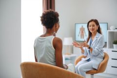 A doctor consults with a patient in a bright, modern office. The doctor, wearing a white coat, is speaking with the patient who is seated in a chair. The scene suggests a medical consultation or therapy session.