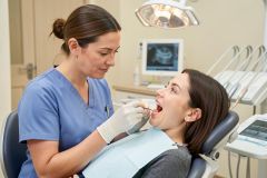 Dentist in scrubs examining a reclining patient's open mouth during an oral exam under the overhead light.