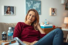 A woman is sitting on the couch in the living room. She is wearing a burgundy top, a water bottle is on the table in front of her and she looks relaxed.