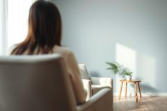 Woman seated in a light, neutral-toned room. Soft sunlight streams across the wooden floor and illuminates a potted plant on a small side table. The scene evokes a calm, peaceful atmosphere, possibly a therapy or waiting room setting. Beige and light blue colors dominate the space.