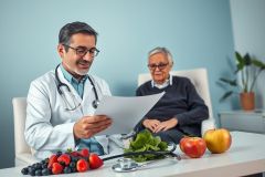 Doctor reviewing patient records in a bright, modern clinic setting. An elderly patient sits nearby. Fresh fruits and vegetables like berries, apples, and spinach are displayed on a table, suggesting a focus on healthy eating and preventative care. Blue and white color scheme.