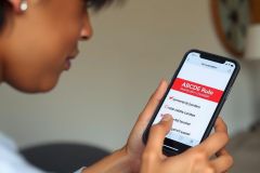 A person is seen holding a smartphone in their hands. The screen displays an app with a red header that has the word 'Rules' prominently visible.