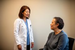 Female clinician in a white coat speaking reassuringly with a seated middle-aged patient in an exam room.
