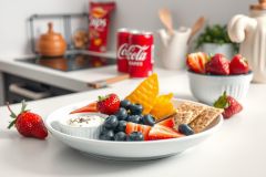 Healthy snack featuring berries, yogurt, and fruit slices arranged on a white plate. The scene includes a bright kitchen with visible food and drinks, suggesting a wholesome lifestyle.