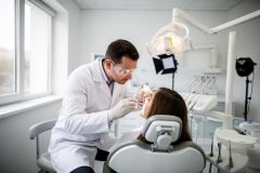 Dentist in protective eyewear examines a patient's teeth with a mirror and probe in a bright clinic.
