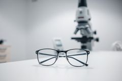 Close-up of eyeglasses on a white lab table with a blurred microscope in the background. A science or research theme, featuring black frames and neutral colors, suggests laboratory work or optical equipment.