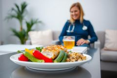 Healthy meal on a plate featuring grilled fish, vegetables, and rice. A woman in a medical uniform sits in the background. Focus on wellness and nutrition.