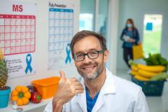 The image shows a man in a lab coat smiling and giving a thumbs up gesture. He is surrounded by various health-related items such as fruits, vegetables, and medical equipment. The environment suggests that he is a scientist or researcher working on health topics.