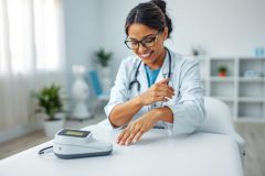 A smiling female doctor in a white coat and glasses examines a patient's hand near a blood pressure monitor in a bright, modern medical office. Healthcare, wellness, and medical examination themes are present.