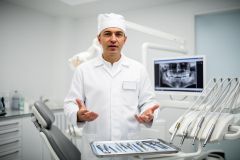 Dentist in a clinical operatory explaining dental instruments, with a panoramic dental X-ray on the monitor.