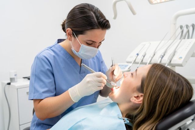 Dental clinician in mask and gloves examines a patient's open mouth with a mirror and probe in a clinic chair.