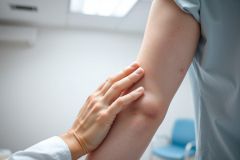 Close-up of a healthcare professional examining a patient's arm in a clinical setting. The image shows a hand gently touching the arm, likely assessing skin condition. Soft, neutral colors and a medical environment suggest a dermatology or wellness checkup.
