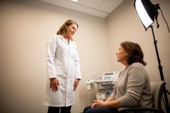 Doctor in a white coat talking with a seated patient in a clinical exam room during a consultation.