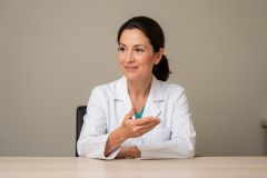 Smiling dental clinician in a white coat sits at a desk, gesturing while discussing treatment options.