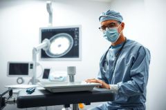 Surgeon in scrubs and mask, seated at an operating table with medical equipment. Sterile environment, blue and gray tones. Healthcare professional, surgical procedure, hospital setting.