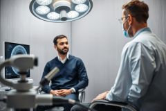 Doctor consultation in a modern medical office. A man discusses health concerns with a doctor wearing a face mask, with medical equipment and a monitor. Blue and gray tones dominate the clinical setting.
