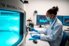 A scientist in a lab coat and gloves analyzes data on computer monitors near a scientific instrument. The scene is dominated by blue and white tones, evoking a biotechnology or medical research environment.
