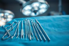 Surgical instruments laid out on a blue operating table under bright surgical lights. Close-up view of stainless steel tools including forceps, scissors, and probes, creating a sterile medical environment. Focus on healthcare, surgery, and medical equipment.