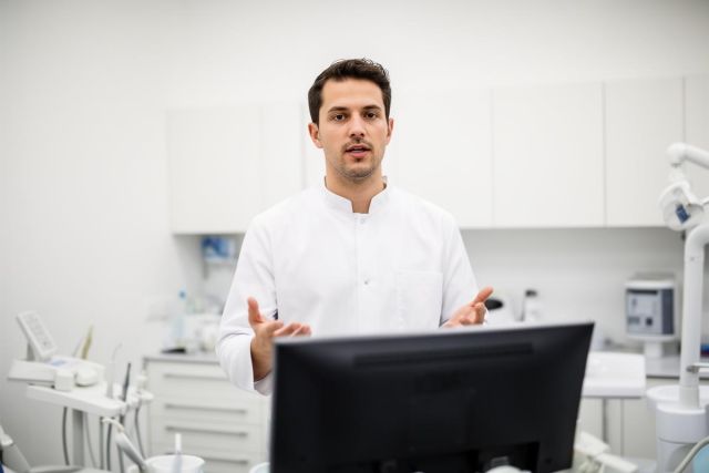 Young surgeon in a white coat speaking across a computer monitor in a clinical exam room, gesturing with hands.