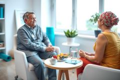 Two people sit and converse in a bright, modern living room. Warm sunlight streams in, illuminating the cozy space with comfortable seating and a round table with fruit. The atmosphere is relaxed and friendly.