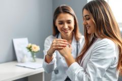 Two female doctors in white coats hold hands in a supportive medical consultation. The scene takes place in a modern, light-filled office setting with soft colors. Focuses on healthcare, empathy, and doctor-patient connection.