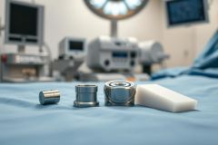 Close-up of surgical tools and components on a blue operating table, with medical equipment blurred in the background. Features include stainless steel parts and a white surgical wedge, suggesting an orthopedic or surgical procedure. Clean, sterile environment with cool blue and white tones.