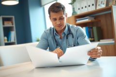 Doctor reviewing patient records at a desk in a medical office. Wearing a blue shirt and stethoscope, he focuses on paperwork in a brightly lit environment.
