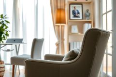 Modern living room with gray armchairs, a bookshelf, and a laptop on a table. Bright, airy space with natural light and a minimalist aesthetic. Features a portrait and indoor plants.