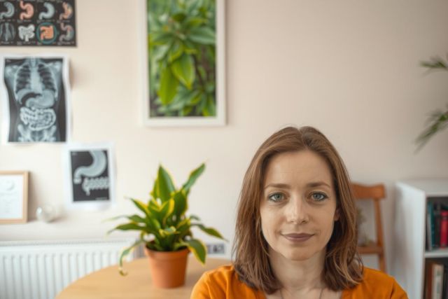 The image features a woman seated at an indoor setting, presumably a home or office. She is wearing an orange top and has a slight smile on her face. 