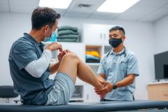 Doctor examining a patient's ankle injury in a medical office. The patient is seated on an examination table, and the doctor is wearing a medical uniform. The scene highlights healthcare and physical therapy.