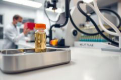 Lab equipment including a vial of liquid on a metal tray. Science laboratory setting with researcher in background. Focus on the vial and laboratory environment. Bright, clean, and professional setting.