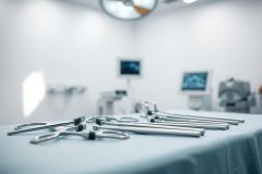 Surgical instruments laid out on a blue surgical table in a blurred operating room setting. Stainless steel tools are visible under bright overhead lighting, suggesting a medical or healthcare context. Clean, sterile environment with monitors in the background.