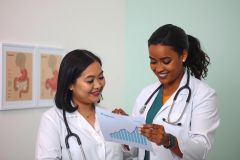 Two doctors in white coats examine a chart in a medical office setting. The scene highlights healthcare professionals reviewing patient data in a clinical environment, featuring a green wall and medical diagrams.