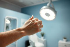 Close-up of a hand with a red ring under bright medical examination room lighting. Blue walls and a blurred dentist chair are visible in the background, suggesting a dental or healthcare setting. Focus on hand and light.