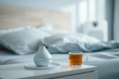 Relaxing bedroom scene featuring an ultrasonic diffuser releasing mist and a cup of tea on a white nightstand. Soft blue and white tones create a calming atmosphere, perfect for aromatherapy and wellness. Cozy and peaceful interior design.