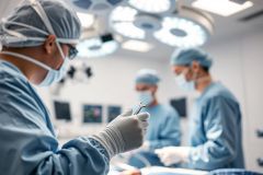 Surgical team performing a medical procedure in a sterile operating room. Focus on gloved hands holding surgical instruments under bright overhead lights. Blue and white color palette, emphasizing a clean and professional healthcare environment. Represents surgery, medical professionals, and hospital setting.