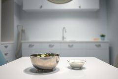 Modern kitchen interior featuring a stainless steel bowl with salad and a small white bowl on a white countertop. The scene showcases a light blue kitchen with cabinetry and a sink, creating a clean and bright culinary space. Food preparation and kitchen design elements are visible.