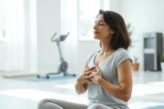 Woman meditating in a bright, modern home gym. She's seated in a relaxed pose, practicing mindfulness with eyes closed. Light gray and white tones dominate the scene, conveying tranquility and wellness. Focus on yoga, meditation, fitness, and mental health.