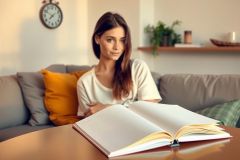 The image shows a woman seated comfortably on a couch. She appears to be engaged in reading or studying. The environment suggests a cozy living room setting with a warm color palette and soft lighting.