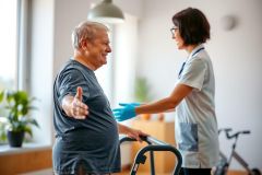 A healthcare worker assists an elderly man with a walker. The scene takes place indoors, suggesting caregiving or rehabilitation.  The man wears a blue shirt, and the caregiver wears a white coat and blue gloves. The setting appears bright and comfortable.
