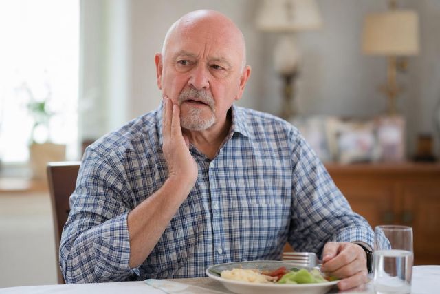 Elderly man at dining table touching cheek, appearing to have dental discomfort while eating.