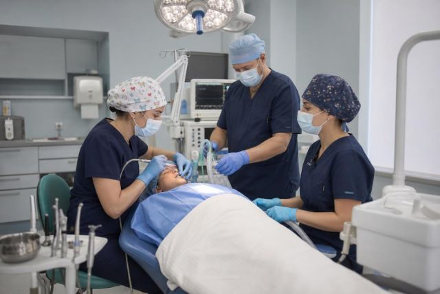 Pediatric dental team administering anesthesia while a child lies on a dental chair monitored.